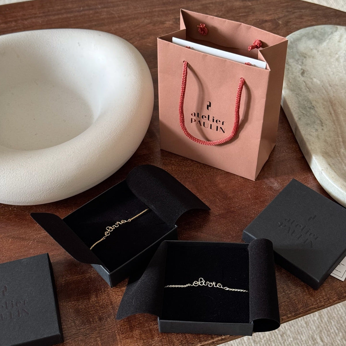 A delicate gold chain bracelet displayed in open black jewellery boxes on a wooden table, alongside a branded Atelier Paulin shopping bag.