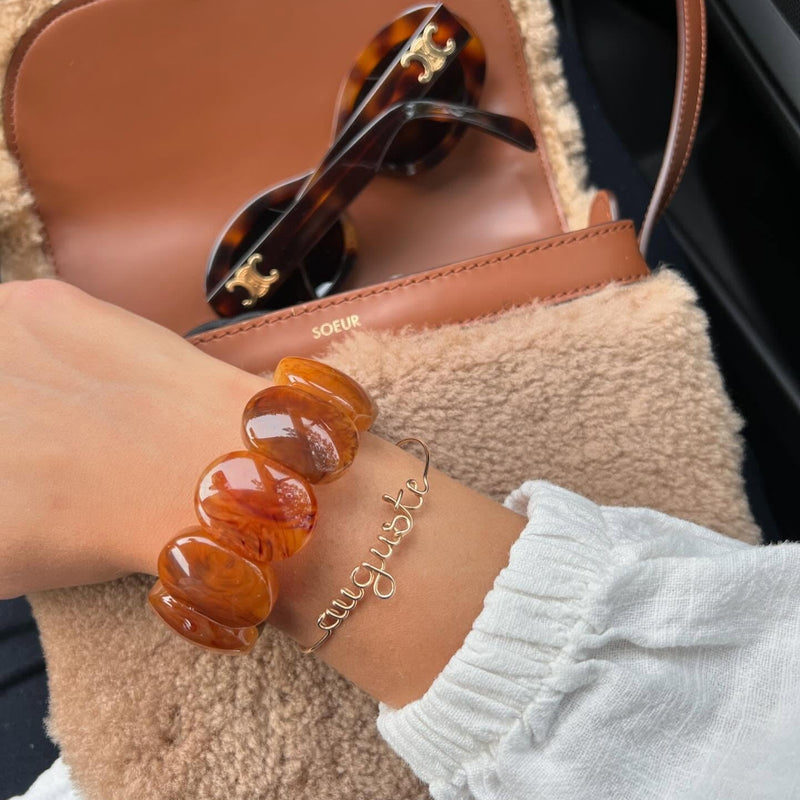Person holding a beige handbag with sunglasses, wearing a gold bracelet with personalised name handwritten.